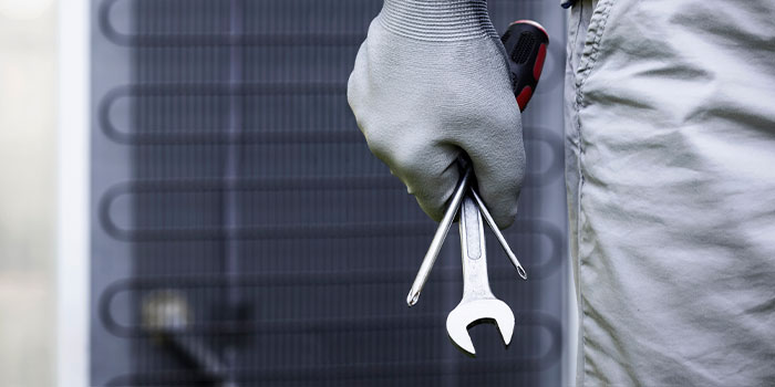 A gloved hand holding wrenches and a screwdriver in front of a cooling unit.