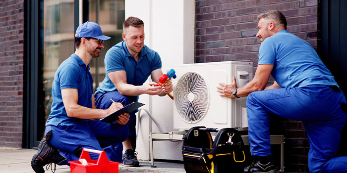Three technicians installing an outdoor air conditioning unit on a building wall.