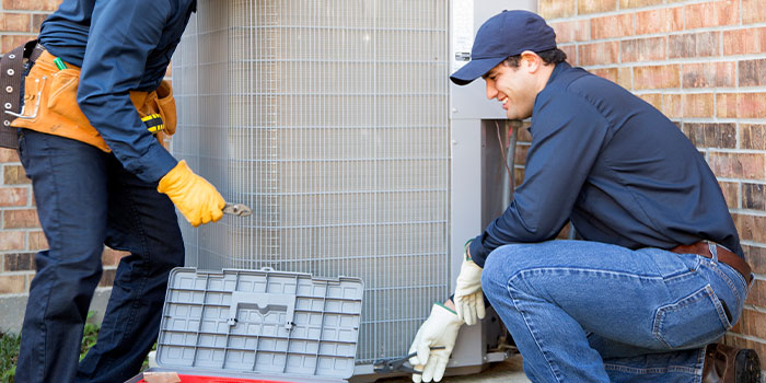 Two technicians servicing an outdoor AC unit with tools.