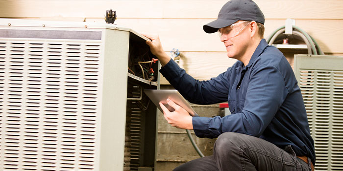 Technician in blue uniform inspecting outdoor HVAC unit with tablet.