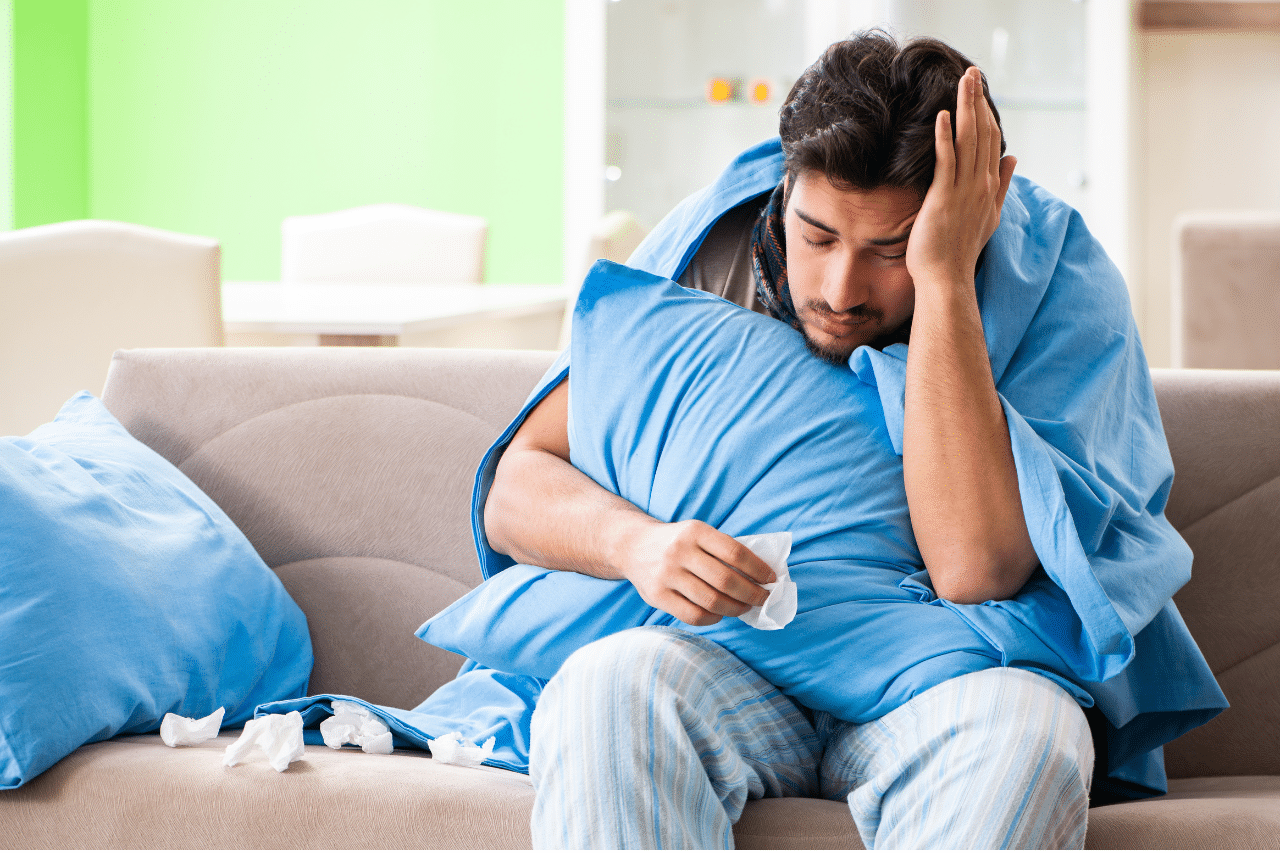 A man wrapped in a blanket sits on a couch looking unwell with tissues around him.