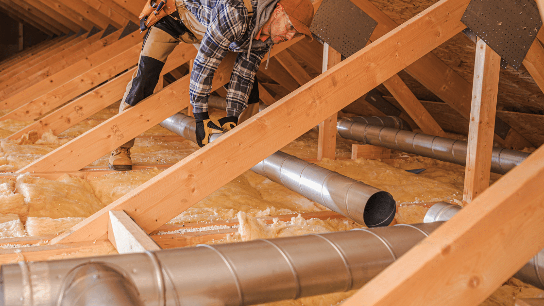 Man installing insulation in an attic, surrounded by wooden beams and HVAC ducts.