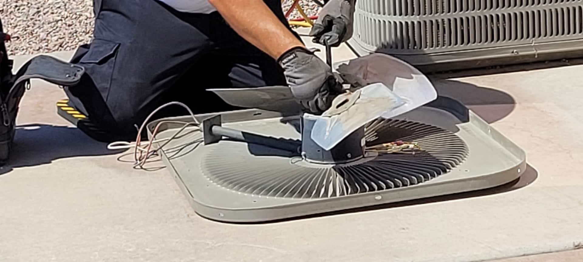 A person repairing an air conditioning unit outdoors, focusing on the fan assembly.