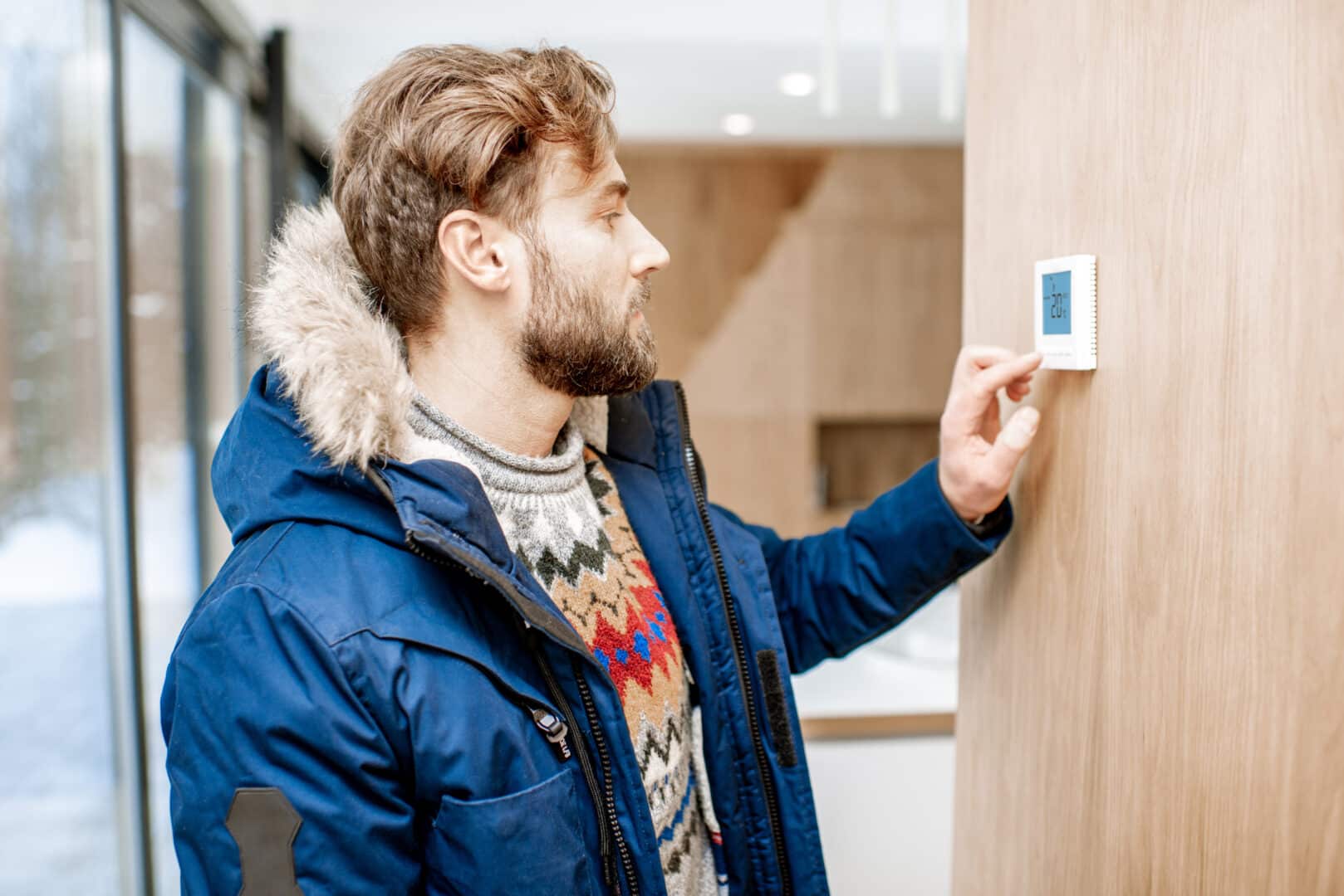 A man in a blue jacket adjusts a thermostat inside a modern home.