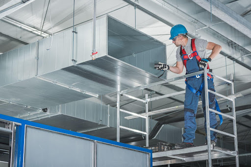 Worker in blue hard hat inspecting air duct system on scaffolding.