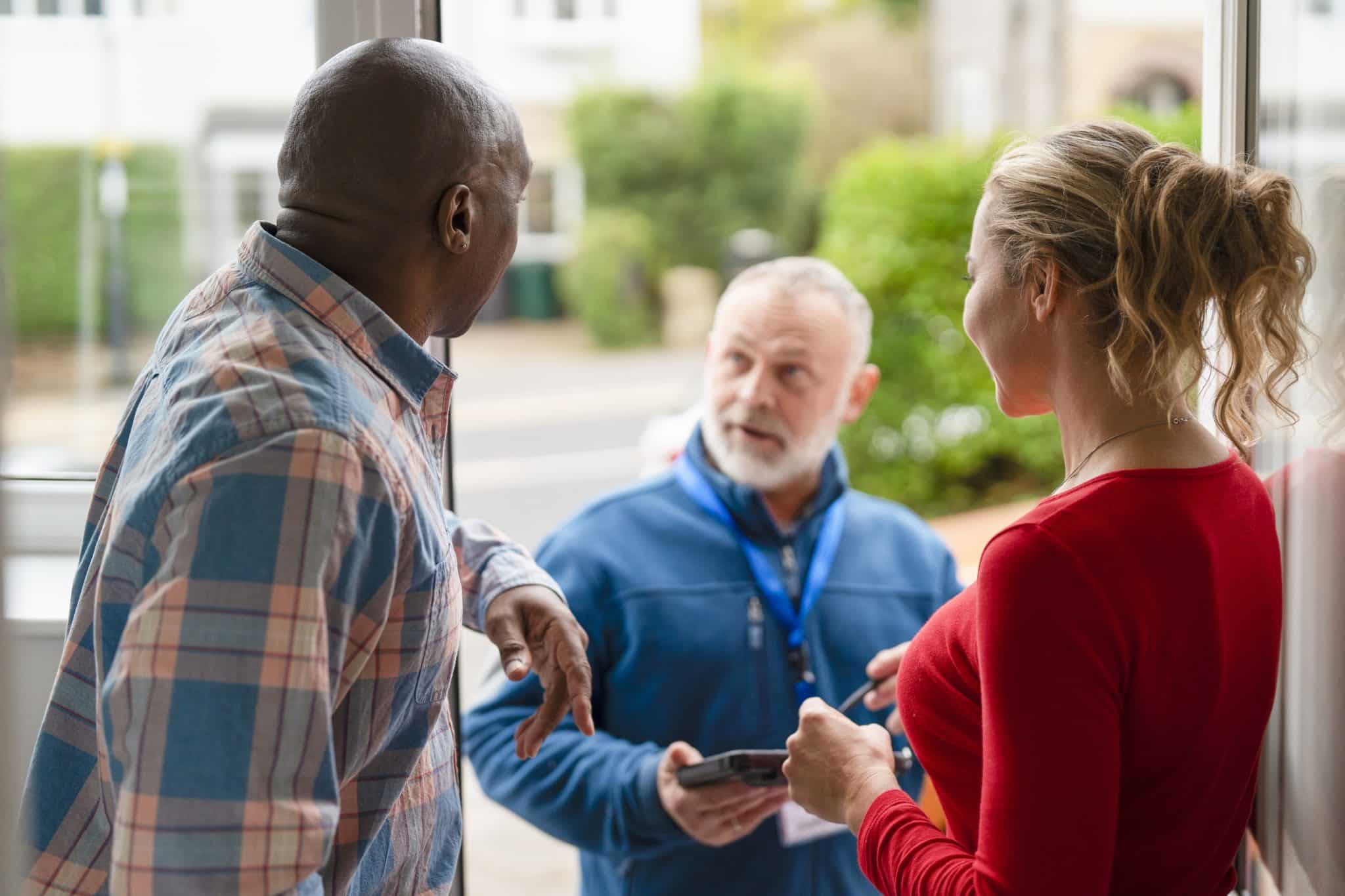 A local HVAC technician chats with homeowners at a doorstep, phone in hand, listening and discussing service needs.